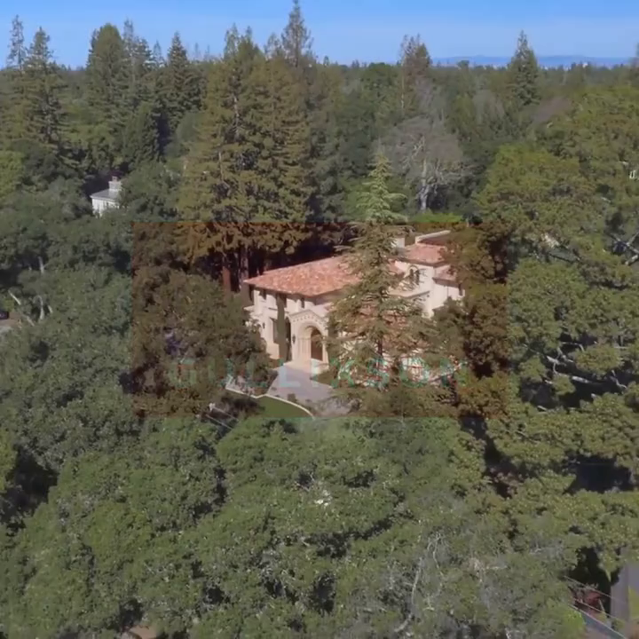 a large beige villa with a red-tiled roof and arched porch, nestled in lush green forest with a manicured lawn and driveway